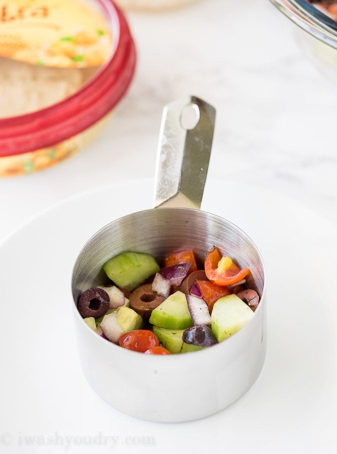 To make these fun Mediterranean Hummus Stacks, simply start by layering your Greek salad in the bottom of a measuring cup.