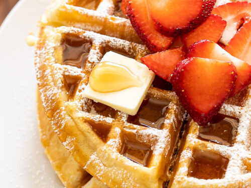 Crispy waffles on white plate with syrup and strawberries.