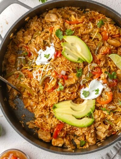 large black skillet filled with fajita rice, chicken and peppers. Topped with cheese and avocado slices.