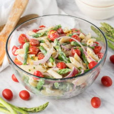 A glass bowl of food on a table, with pasta, asparagus, tomatoes and onions