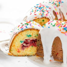A close up of a white frosted bunt cake topped with sprinkles, with a slice slightly removed to show the center of cherry