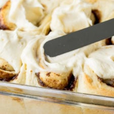 A close up of cinnamon rolls in a pan, being topped with a cream frosting with spatula