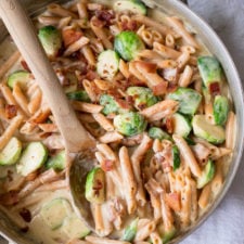 A close up of a pan with a wooden serving spoon scooping food from it, with  pasta, creamy red sauce and veggies