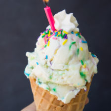 A close up of a waffle cone with a scoop of Creamy Cake Batter Ice Cream topped with whipped cream and sprinkles