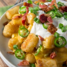 A close up of a plate of Jalapeño Popper Totchos topped with sour cream, bacon and jalapeño slices