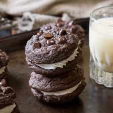 Two chocolate chocolate chip sandwich cookies stacked on top of eachother with a white filling and a side of a glass of milk