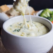 A close up of a piece of bread being dipped into a bowl of spinach and artichoke fondue