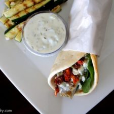 A wrapped Grilled Chicken Souvlaki Pita on a plate next to a small bowl of dipping sauce and grilled zucchini strips
