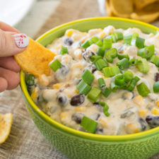 A person holding dipping a chip into a white cream dip with corn, black beans and topped with chives