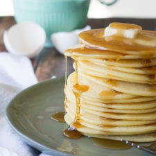 A large stack of thin pancakes on a plate topped with drizzling syrup and a square of melting butter
