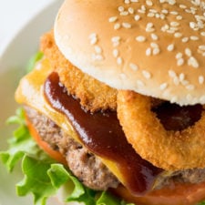 A close up of a hamburger on a plate with cheese, bbq sauce and onion rings