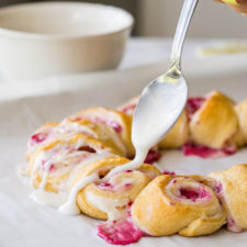 A plate of food with lemon raspberry danish rolls being topped with a frosting