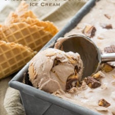 A close up of ice cream with peanut butter cups being scooped from a pan and cones next to the pan on the table