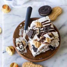 A slice of ice cream cake on a plate with cookies and chocolate sauce and peanut butter cookies, Oreos and peanut butter cups surrounding it