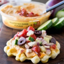 A close up of a waffle topped with hummus, olives, tomatoes, zucchini and cheese