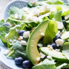 A close up of a bowl of salad, with spinach, avacado, blueberries and feta cheese