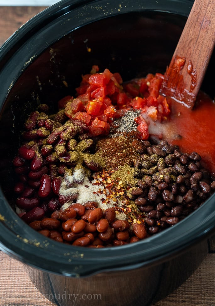 canned beans with ground beef and spices in slow cooker, ready to mix with wooden spoon.