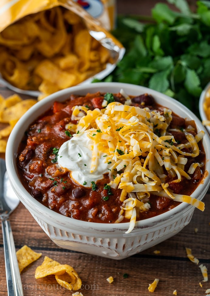 thick and hearty chili in white bowl with sour cream and shredded cheese on top.