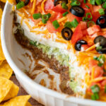 7 Layer Bean Dip with scoop taken out to show layers of beans, guacamole, sour cream and cheese.