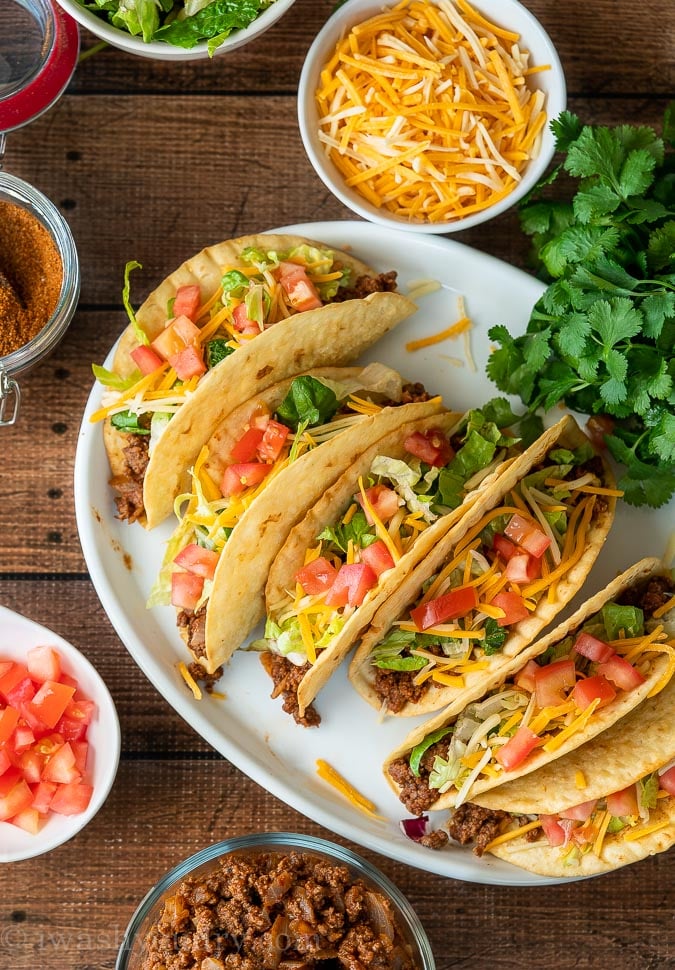 A plate full of crispy flour tortilla ground beef tacos with toppings.