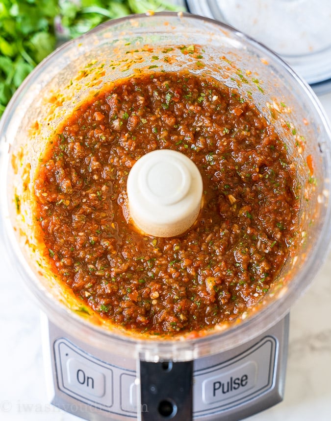 Blended salsa ingredients shown in the food processor, bright red color with flecks of green cilantro throughout.