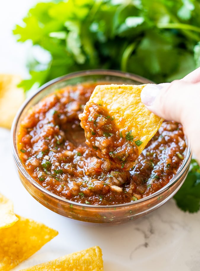 A small bowl filled with delicious salsa and a hand dipping a yellow corn tortilla chip into it.