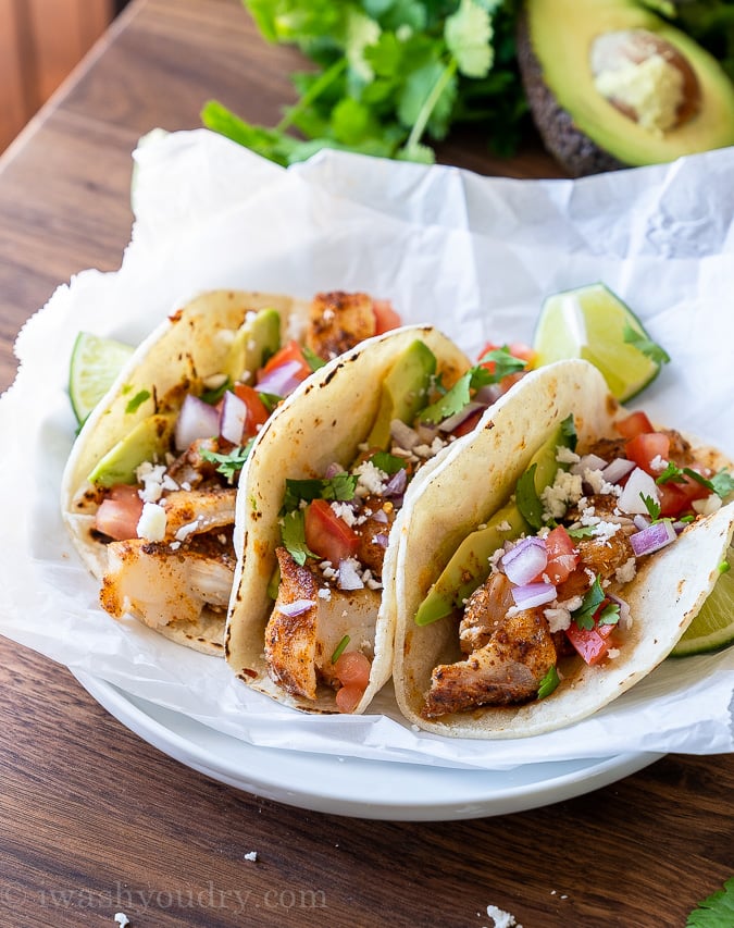 Three fish tacos on a plate, topped with avocado, diced tomato, onion and cilantro.