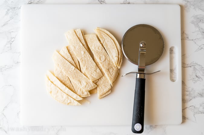 Strips of white corn tortillas with a pizza cutter beside them on a white cutting board.