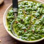 Small white bowl filled with green chimichurri sauce and red pepper flakes.