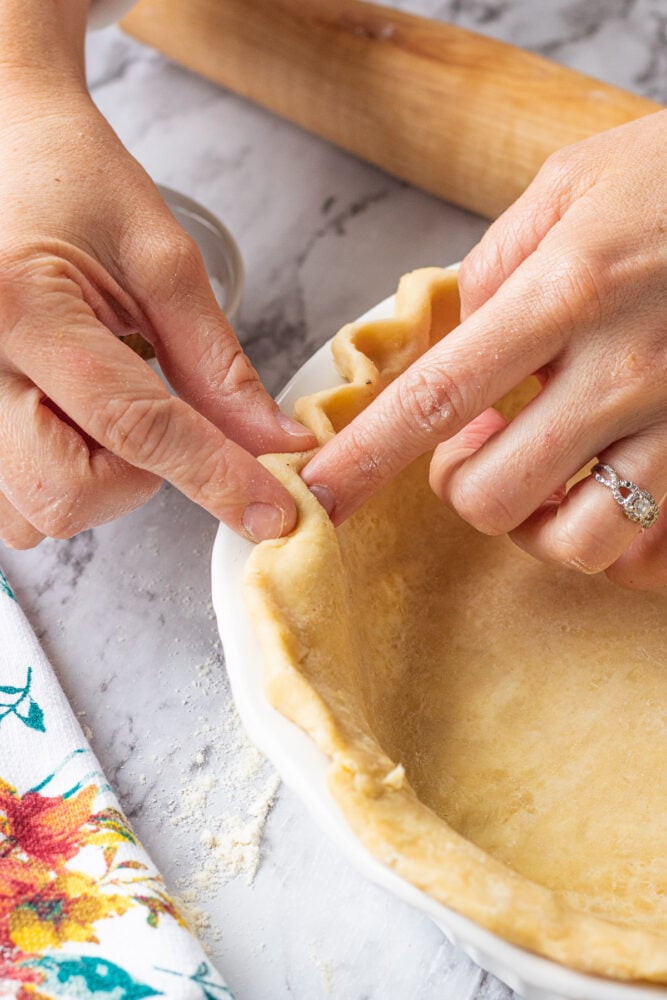Hands crimping raw pie dough in white pan. 