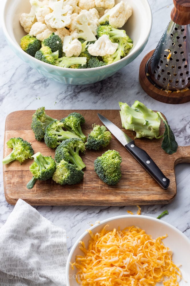 Chopped broccoli on wood cutting board with knife, by bowl of broccoli and cheese grater on marble. 