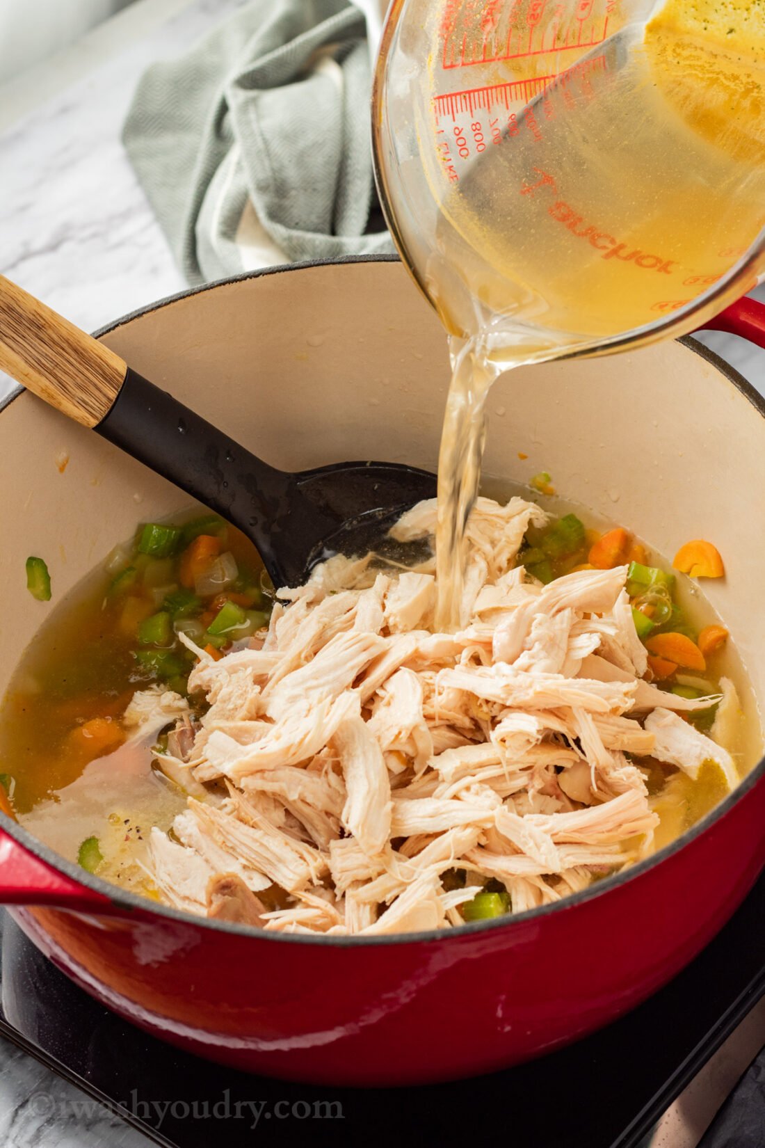 Pouring chicken broth on top of rotisserie chicken and cooked carrots, celery, and onion in red pot. 