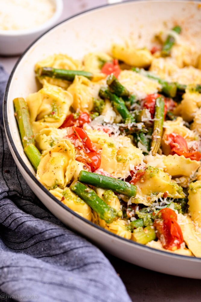 Cooked tortellini, asparagus, and tomatoes in white skillet.
