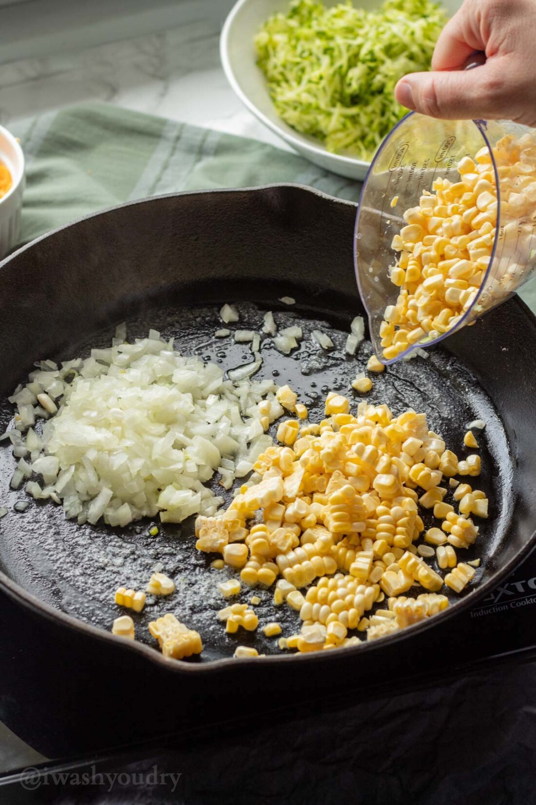Pouring corn into black frying pan with chopped onion. 