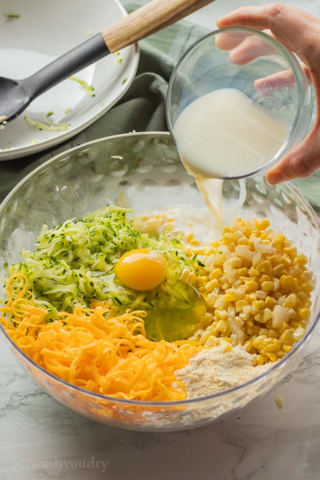 Pouring milk and egg into bowl of cheese, zucchini, corn, onion, flour, cornmeal, and spices. 