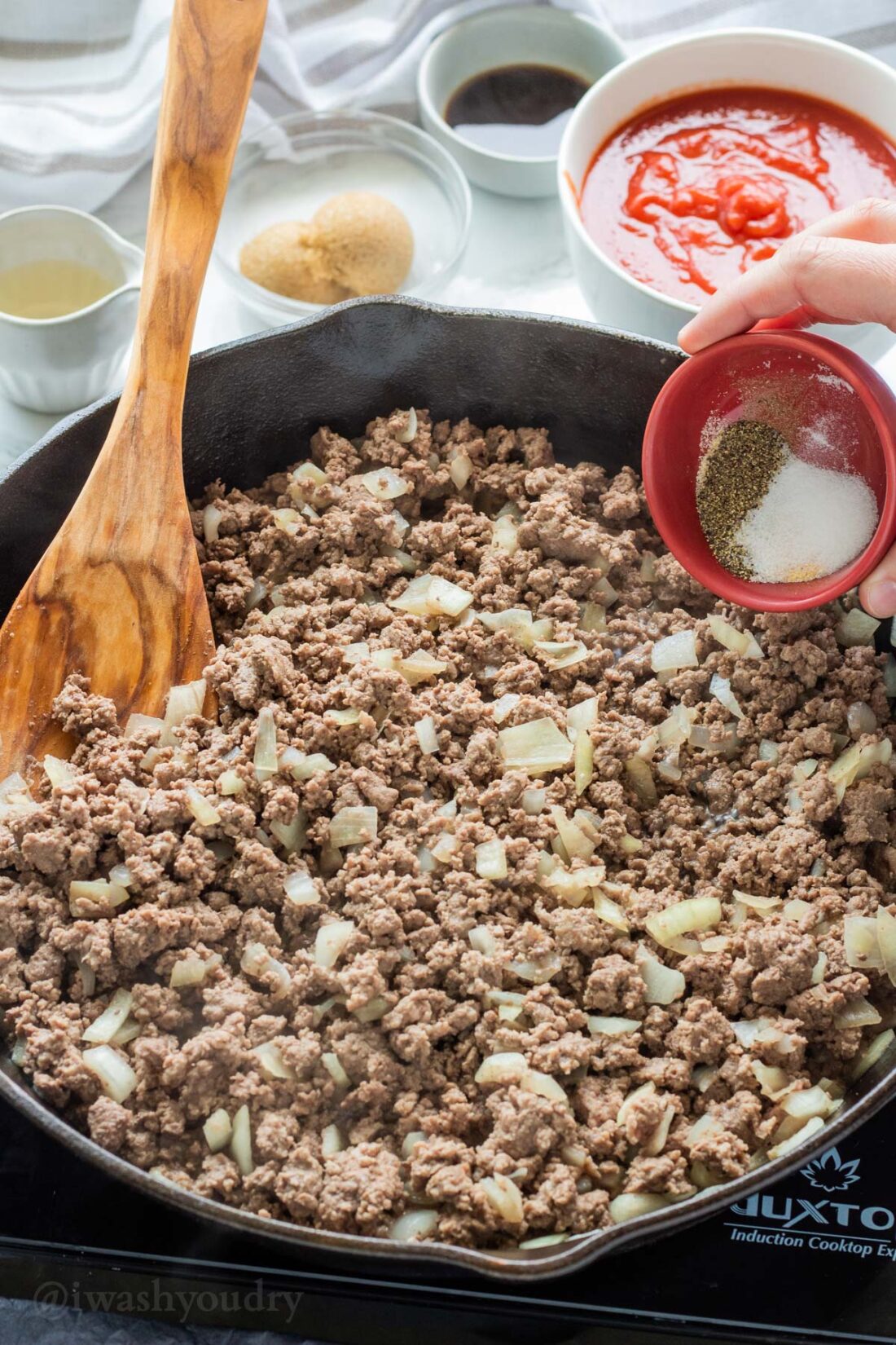 Pouring a red bowl of spice into a black frying pan of cooked ground beef. 