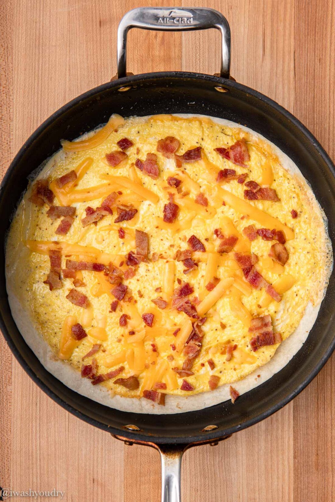 Cooked eggs, cheese and bacon on tortilla in a frying pan.
