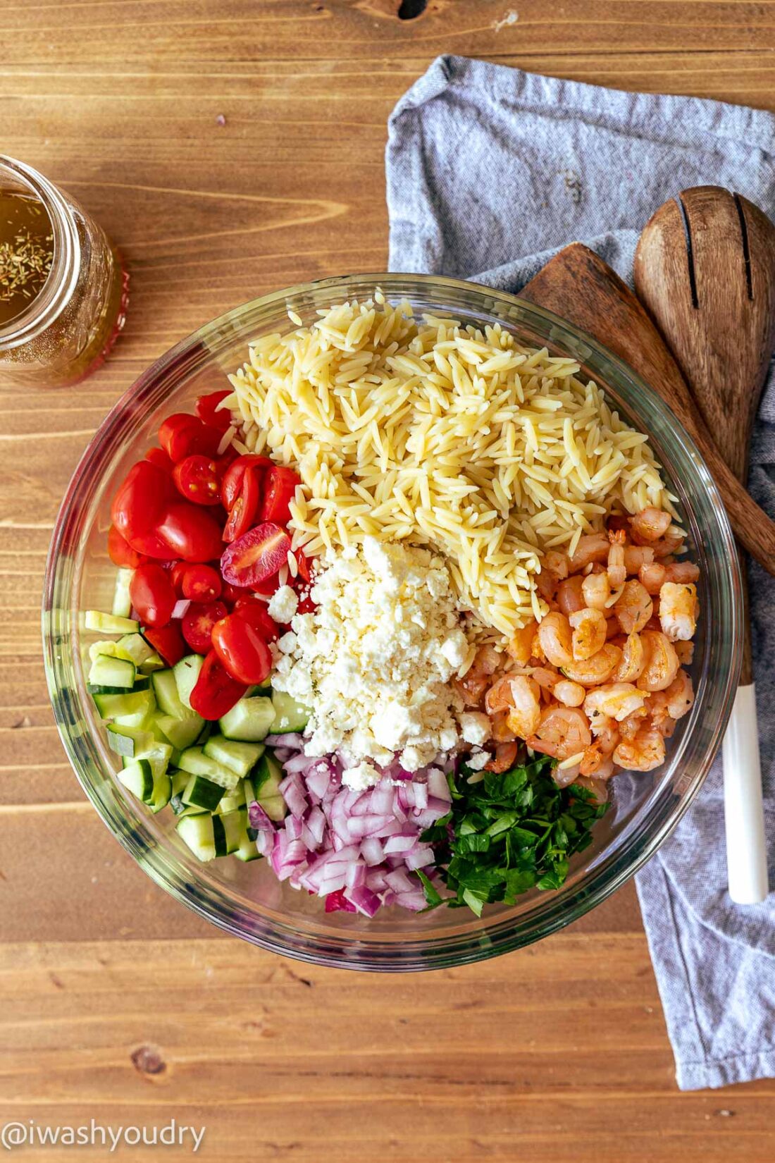 Shrimp orzo pasta salad ingredients in glass bow with wooden spoonsl on wood cutting board. 