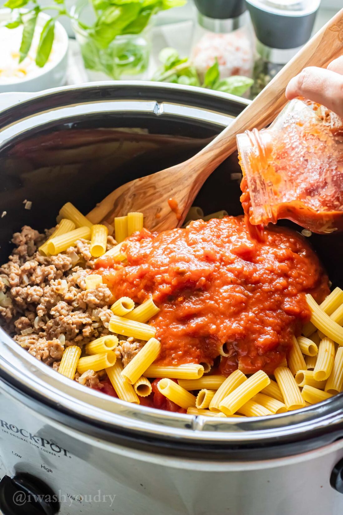 Pouring marinara sauce into slow cooker of dry ziti noodles and cooked italian sausage. 