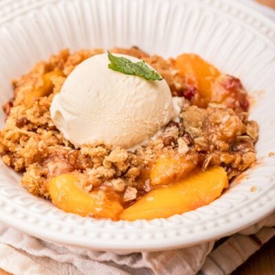 Baked peach cristp in a white bowl on a tea towel.
