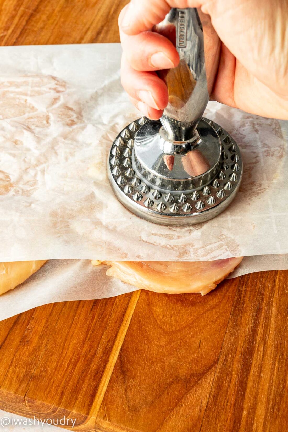 Hand using meat tenderizer on raw chicken breast in parchment paper. 
