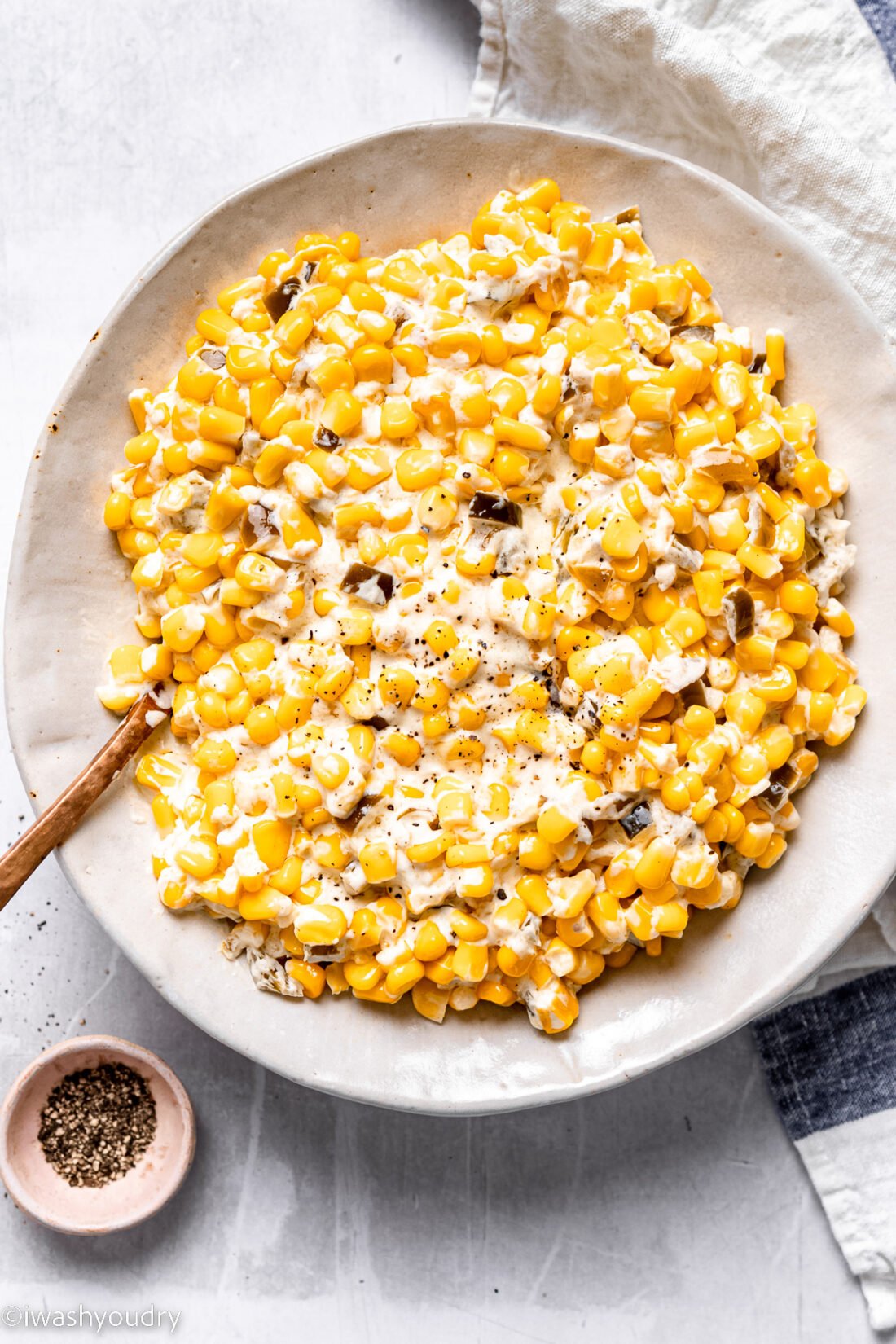Cooked slow cooker creamed corn in a white bowl with small bowl of pepper. 