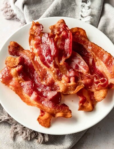 Cooked bacon slices on white plate with dish towel.