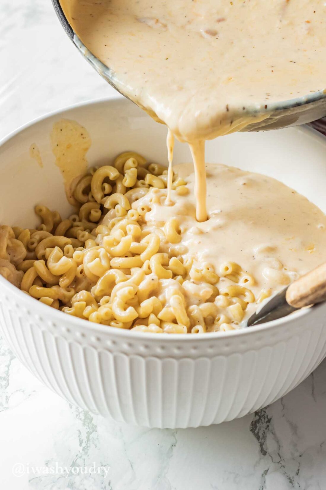 Pouring cheese sauce into cooked macaroni noodles in a white bowl. 