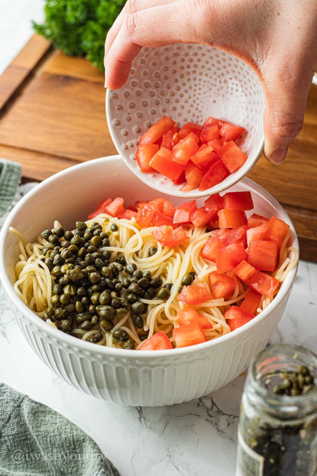 Hand pouring white bowl of tomatoes into larger bowl of noodles and capers. 