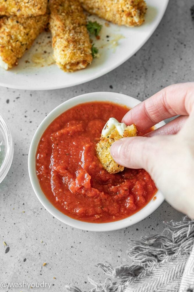 Dipping mozzarella sticks into marinara sauce in white bowl.