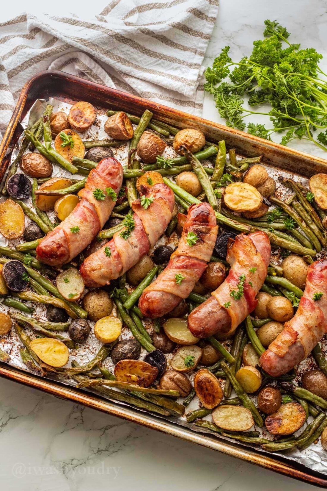 Cooked bacon wrapped bratwurst on roasted vegetables in metal baking pan.