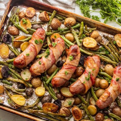 Cooked bacon wrapped bratwurst on roasted vegetables in metal baking pan.