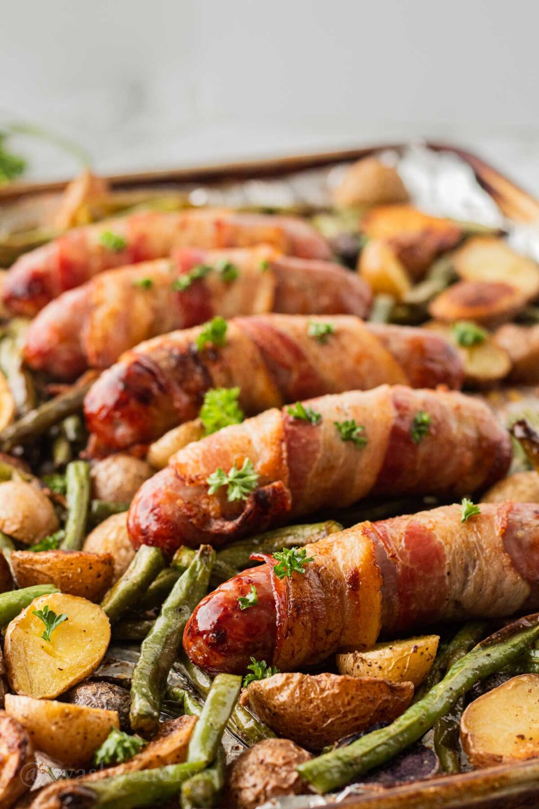 Cooked bacon wrapped bratwurst on roasted vegetables on metal sheet pan. 