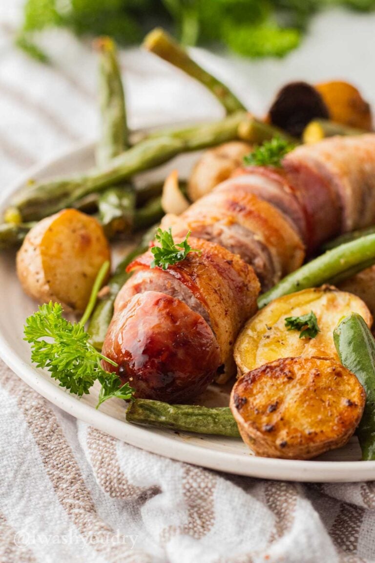 Cooked and sliced bacon wrapped bratwurst with potatoes and green beans on white plate.